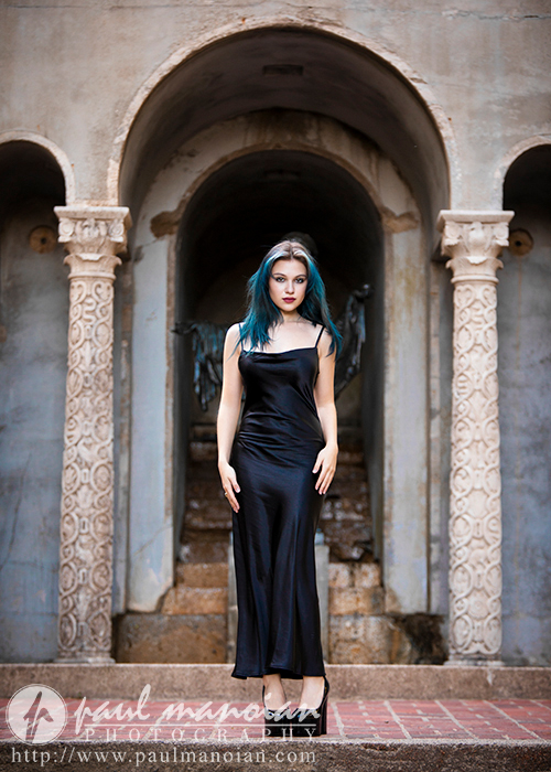 A high school senior with blue hair and a long black dress stands confidently in an archway surrounded by ornate stone pillars. The backdrop features weathered stone steps and an aged wall. The photo credit reads "Paul Manoian Photography" with a web address below.