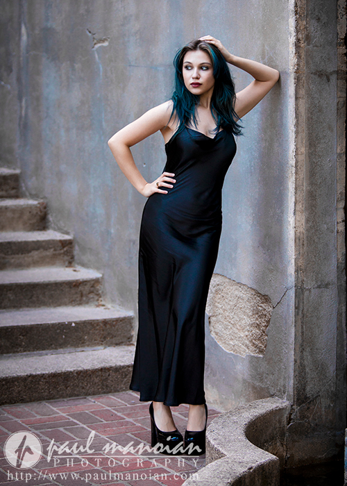 A high school senior with blue-green hair stands against a worn stone wall with her right arm resting above her head. She is wearing a long, black sleeveless dress and high-heeled shoes. Stairs are visible to her left. The photo is watermarked with the photographer's logo and website.