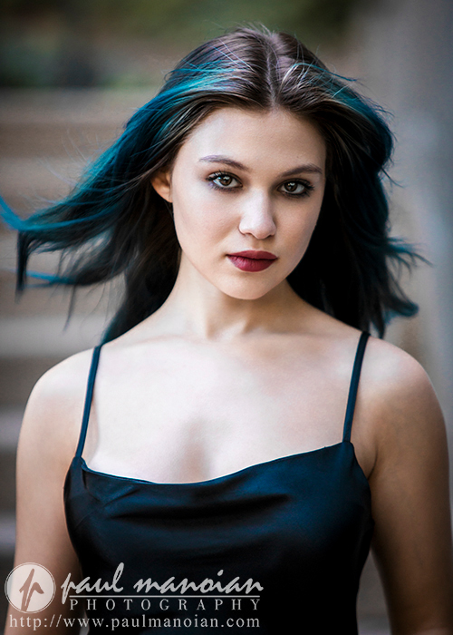 A high school senior with long, slightly wind-swept dark hair with blue highlights, wearing a black spaghetti-strap top, stares directly at the camera. The background is blurred, highlighting her strong gaze. The bottom of the image features the text "paul manoian PHOTOGRAPHY" and a website link.