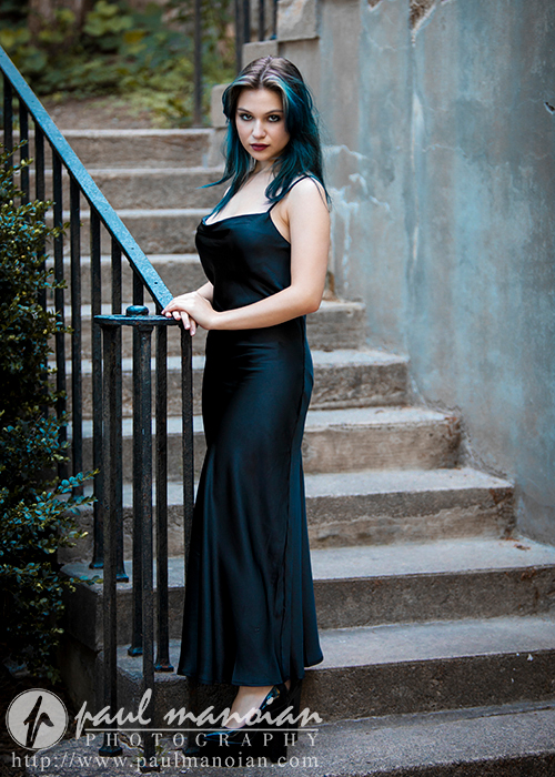 A high school senior with teal hair is standing on an outdoor staircase, leaning on the railing. She is wearing a long, sleeveless black dress. There is greenery beside her and textured, concrete walls around the staircase. The image is marked with a photography logo and website URL.
