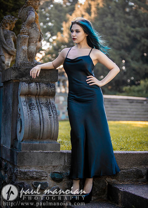 A high school senior in a dark blue dress stands by a stone structure adorned with sculptures in an outdoor setting. Her hair is blue, and she gazes thoughtfully into the distance with one hand resting on the structure. The setting is lush, with trees and steps in the background.