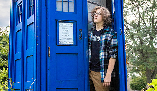 A person with long hair, wearing glasses, a black t-shirt, a plaid shirt, and tan pants stands partially inside an open, blue, police box in a garden. Trees and cloudy sky are in the background.