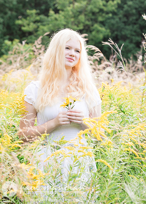 A high school senior girl named Alyvia with long, blonde hair stands in a field of tall yellow wildflowers in Livonia. She is wearing a white dress and holding a small bunch of flowers. The background is lush and green with trees, and the sunlight illuminates her face and hair, perfect for senior portraits.