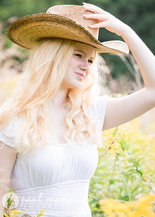 A high school senior with long blonde hair wearing a white dress and a large straw hat stands in a field of yellow flowers. She has a slight smile and looks into the distance, lightly touching the brim of her hat with one hand. Capturing the essence of Livonia's charm, it's an ideal scene for Alyvia's senior portraits.