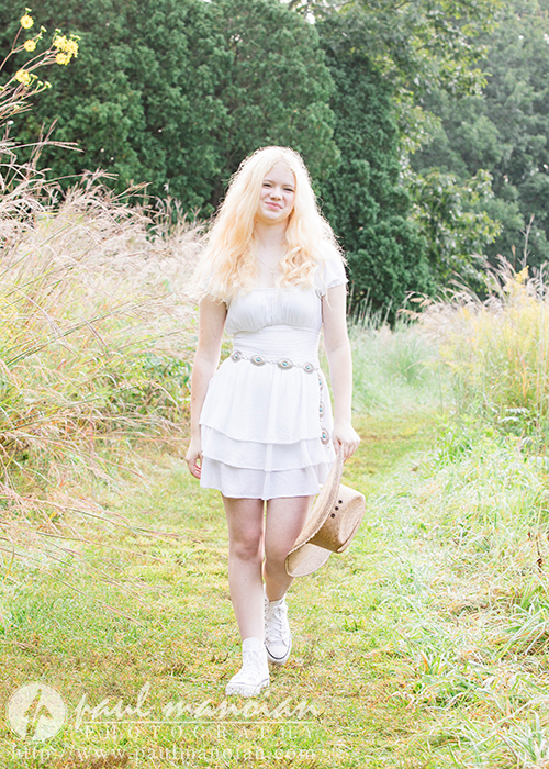 A high school senior firl with long blonde hair, wearing a white dress and white sneakers, walks along a grassy path through tall wild grasses. Alyvia holds a straw hat by her side, with a slight smile on her face, and tall trees can be seen in the background—a perfect scene for Livonia Senior Portraits.