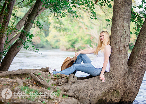 Alyvia, a blonde high school senior in a white top and blue jeans, sits on a large tree branch by a river, holding a brown hat. The background features lush green foliage and the flowing river. Livonia Senior Portraits captured this serene moment. Photo credit: Paul Manoian Photography.