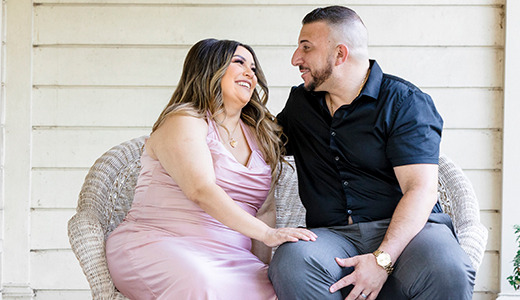 A couple sits on a wicker chair outside, smiling and looking at each other during a maternity photoshoot. The woman wears a pink dress, and the man wears a black shirt and gray pants. They are sitting close together on a porch with light-colored siding and columns.