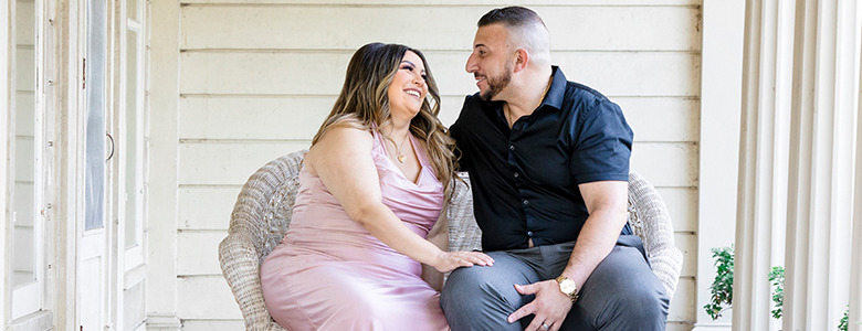 A couple sits on a wicker chair outside, smiling and looking at each other during a maternity photoshoot. The woman wears a pink dress, and the man wears a black shirt and gray pants. They are sitting close together on a porch with light-colored siding and columns.