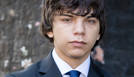 A young man with medium-length, dark hair standing in front of a blurred brick wall. He is wearing a dark suit and a blue tie, looking directly at the camera with a neutral expression.