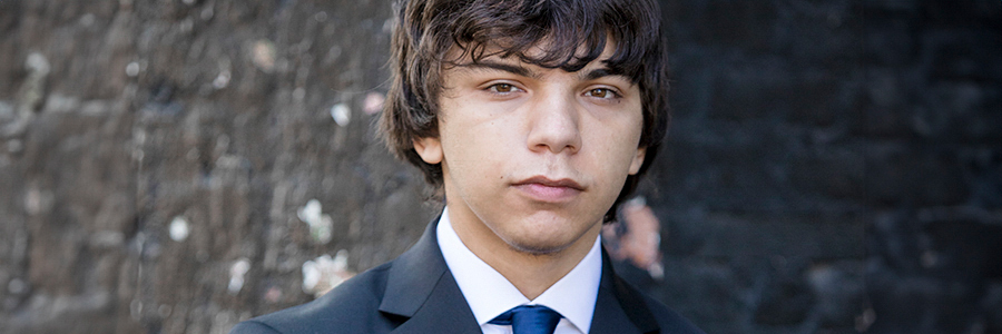 A young man with medium-length, dark hair standing in front of a blurred brick wall. He is wearing a dark suit and a blue tie, looking directly at the camera with a neutral expression.
