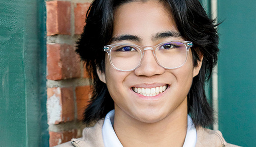 A young person with long dark hair and glasses smiles at the camera. They are standing in front of a brick wall and a green doorframe. They are wearing a light-colored shirt and a beige jacket, captured beautifully in their Rochester Hills senior portraits.