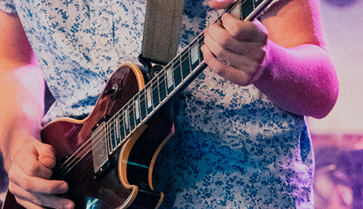 Close-up of a person wearing a short-sleeved floral shirt playing an electric guitar during a live performance. The stage is lit with colorful lights, and a drum set is visible in the background. The image focuses on the musician's torso and arms.