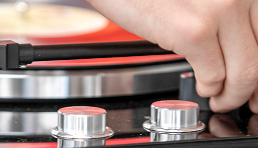 A hand is adjusting the tonearm of a turntable while a vinyl record spins beneath, reminiscent of senior pictures with vinyl records. The image captures the close-up view of the tonearm, knobs, and part of the record player in a soft-focus background.