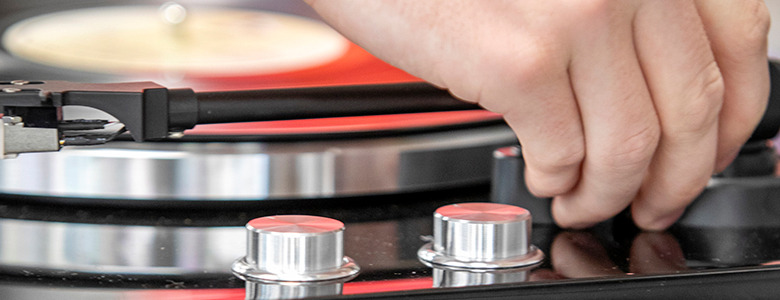 A hand is adjusting the tonearm of a turntable while a vinyl record spins beneath, reminiscent of senior pictures with vinyl records. The image captures the close-up view of the tonearm, knobs, and part of the record player in a soft-focus background.
