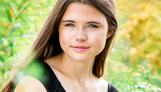 A high school senior girl, capturing her Ann Arbor senior pictures, poses with long brown hair and a black top against a lush, green natural background adorned with yellow flowers. She smiles softly at the camera.