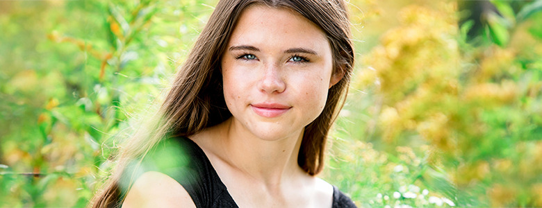 A high school senior girl, capturing her Ann Arbor senior pictures, poses with long brown hair and a black top against a lush, green natural background adorned with yellow flowers. She smiles softly at the camera.