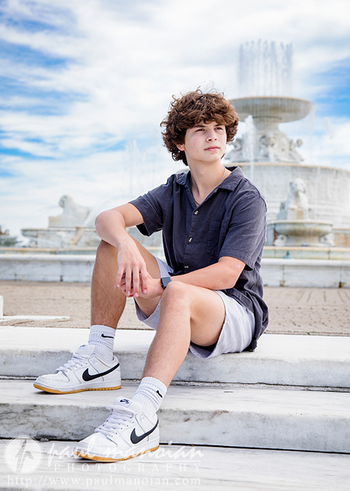 Elijah, a young man with curly hair, sits on a white marble bench in Downtown Detroit. He's dressed in a dark short-sleeve shirt, light shorts, white Nike sneakers, and white socks. A large, ornate fountain is visible behind him under a partly cloudy sky—perfect for his senior portraits.
