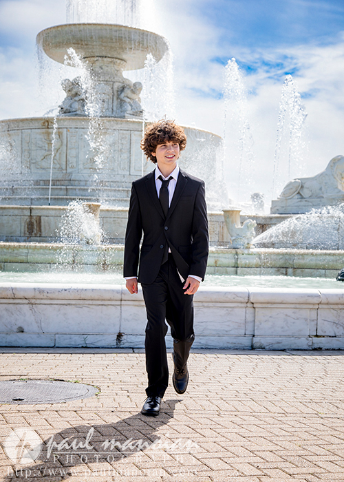 Elijah, dressed in a sleek black suit and tie, walks confidently on a paved path in downtown Detroit. The elegant marble fountain sprays water, creating a majestic backdrop for his senior portraits. With the bright sky above, it's clear this sunny day is meant for capturing moments.