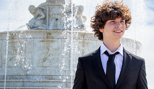 Elijah, a young person with curly hair, dressed in a black suit and white shirt, stands in front of an ornate fountain with water spraying. The sky is blue with some clouds, and statues are visible on the fountain. Downtown Detroit serves as the backdrop for his senior portraits, and he appears to be smiling.