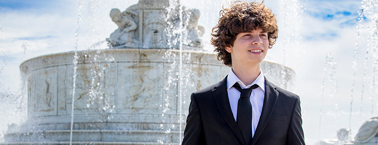 Elijah, a young person with curly hair, dressed in a black suit and white shirt, stands in front of an ornate fountain with water spraying. The sky is blue with some clouds, and statues are visible on the fountain. Downtown Detroit serves as the backdrop for his senior portraits, and he appears to be smiling.