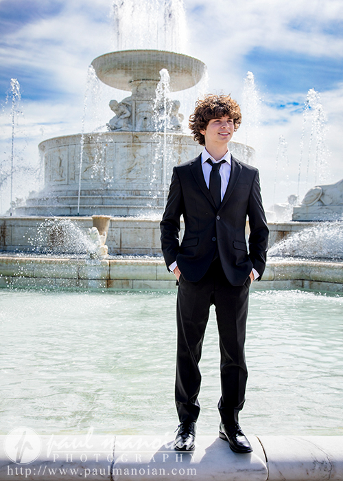 Elijah, a young person with curly hair, stands confidently in front of a large, ornate fountain in Downtown Detroit. They are wearing a dark suit with a white shirt and tie. The sky is blue with some clouds, and water jets are spraying elegantly behind them, making for stunning senior portraits.