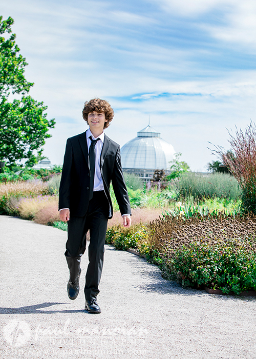 A young man with curly hair, dressed in a black suit and tie, walks along a garden path. The background includes lush green trees, various plants and flowers, and a large domed greenhouse under a blue sky, capturing the elegance of downtown Detroit senior portraits.