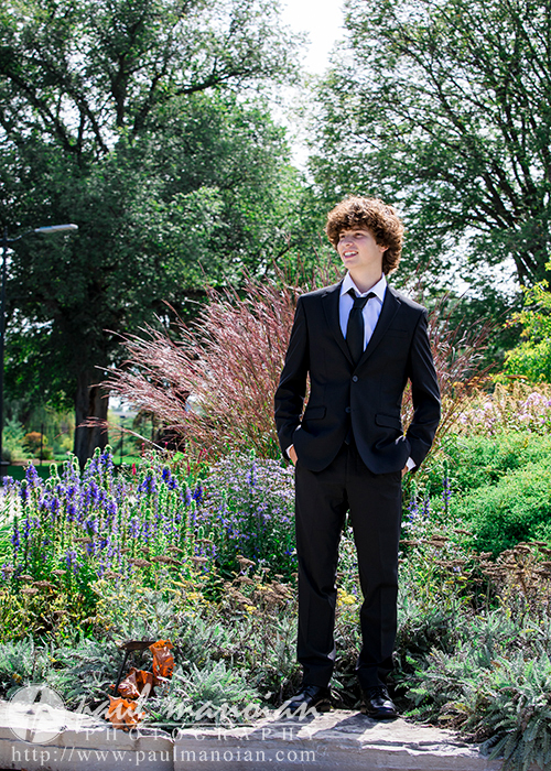 A young person with curly hair stands outdoors in a garden, wearing a black suit and tie, smiling while looking to their left. The background is filled with greenery, colorful flowers, and tall trees. The watermark indicates this is part of downtown Detroit senior portraits.