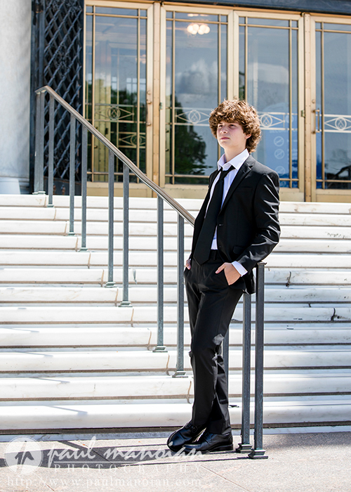A young person with curly hair, dressed in a black suit and tie, stands confidently with one hand in their pocket, leaning against a railing in front of an elegant building with large glass doors and a staircase—classic downtown Detroit senior portraits.