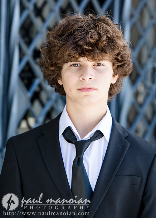 A young person with curly hair stands in front of a textured background, wearing a black suit, white shirt, and a loosely knotted black tie. The photo, part of the downtown Detroit senior portraits collection by "Paul Manoian Photography," includes a watermark with the website URL.