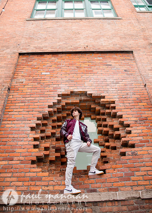 A high school senior wearing a maroon letterman jacket and white pants stands confidently in a circular opening in a brick wall, embodying the urban charm of downtown Detroit. Above them, large green-framed windows are visible, giving the image a distinct vibe perfect for senior portraits.