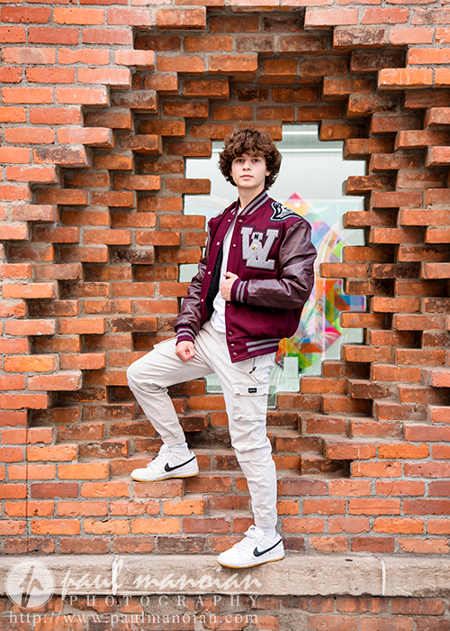 A high school senior wearing a maroon letterman jacket and white pants stands confidently in a circular opening in a brick wall, embodying the urban charm of downtown Detroit. Above them, large green-framed windows are visible, giving the image a distinct vibe perfect for senior portraits.