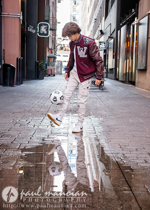 A high school senior wearing a maroon varsity jacket and white sneakers is seen balancing a soccer ball near a puddle in a narrow alleyway, capturing the essence of downtown Detroit senior portraits. The reflection of the person and the ball is visible in the water.