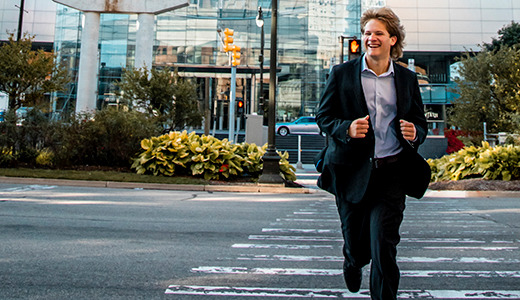 A high school senior in a suit and tie runs confidently across a crosswalk in an urban area. Behind him are modern buildings, greenery, and traffic lights, suggesting a busy city environment. His expression is enthusiastic and he appears to be in a hurry.