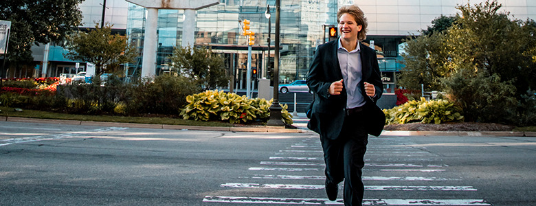 A high school senior in a suit and tie runs confidently across a crosswalk in an urban area. Behind him are modern buildings, greenery, and traffic lights, suggesting a busy city environment. His expression is enthusiastic and he appears to be in a hurry.