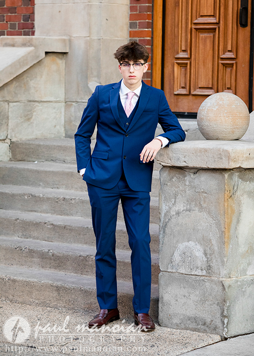 A high school senior in a blue suit stands on a sidewalk next to a stone railing in front of a building entrance, capturing his Urban Senior Pictures. He is wearing glasses, a pink tie, and brown shoes, with his hand resting on the railing and looking confidently at the camera. Steps lead up to a wooden door in Detroit.