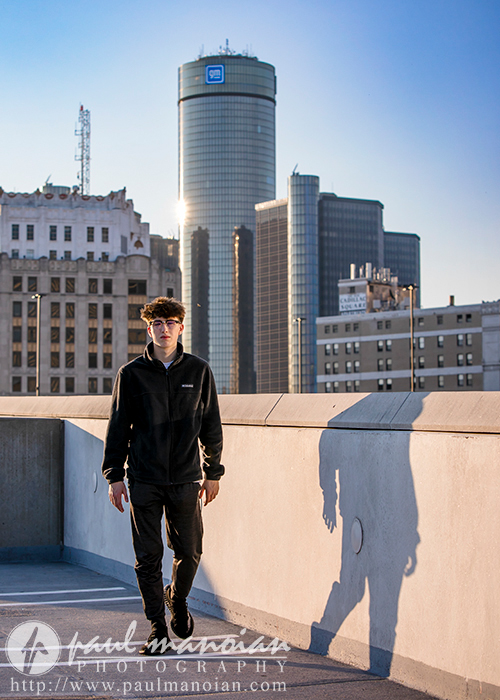 A high school senior wearing a dark outfit and glasses walks on a rooftop parking deck in Detroit. Tall buildings and a large cylindrical skyscraper are in the background, bathed in the warm glow of the setting sun. The person's shadow is cast prominently on the wall, creating an ideal scene for senior pictures.
