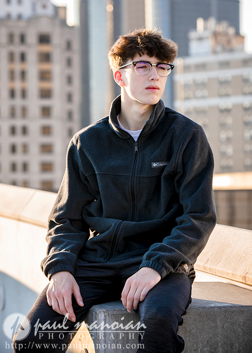 A high school senior with short hair and glasses, wearing a black zip-up jacket, sits on a concrete ledge. They are outside with the urban backdrop of Detroit's cityscape and tall buildings in the background, partially bathed in sunlight.