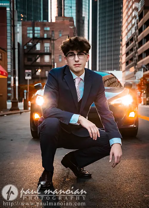 A high school senior wearing a dark suit crouches in the middle of a Detroit city street with tall buildings in the background. The individual is lit by the headlights of a car behind them, creating a dramatic urban scene perfect for senior pictures.