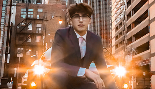 A high school senior in a suit kneels in the middle of a street in an urban area with bright headlights shining behind them, capturing stunning urban senior pictures. Buildings and a Sweet Market bakery are visible on either side of the street, set against the backdrop of dusk or early evening.