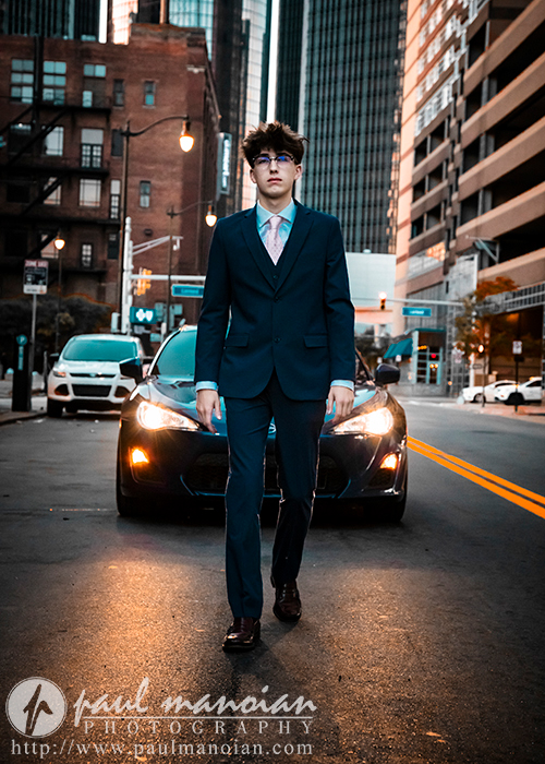 A high school senior with curly hair wearing a suit and tie walks confidently down the middle of a city street, reminiscent of stylish urban senior pictures. It's evening, with cars lining the road behind him, buildings on both sides, and bright headlights illuminating the scene.