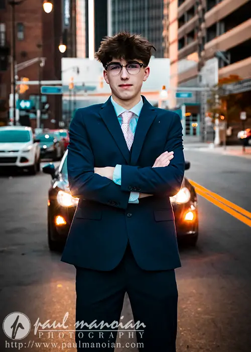 Senior Pictures Outfits Ideas for Guys in 2025: A high school senior with short hair and glasses stands confidently with arms crossed in the middle of a city street, capturing urban senior pictures. The individual is dressed in a dark blue suit with a light-colored tie. Tall buildings and cars are visible in the background, their headlights turned on.