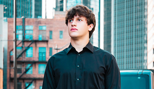 A high school senior with short brown hair in a black button-up shirt stands on a city street in Detroit, with a mix of modern and older brick buildings in the background. He looks off into the distance with a thoughtful expression, capturing the essence of senior portraits.