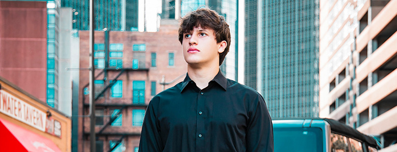 A high school senior with short brown hair in a black button-up shirt stands on a city street in Detroit, with a mix of modern and older brick buildings in the background. He looks off into the distance with a thoughtful expression, capturing the essence of senior portraits.