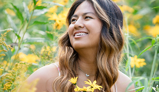 A high school senior with long, wavy hair and braces smiles while surrounded by yellow wildflowers. She is outdoors in a lush, green field, looking slightly to her left. The scene is bright and cheerful, with vibrant flowers adding color to the background.