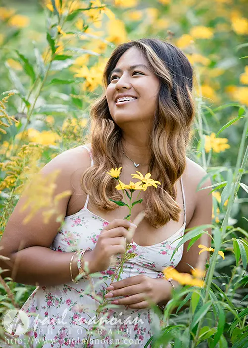 A high school senior with long wavy hair and wearing braces is smiling at the camera. She is dressed in a floral-patterned camisole and standing in front of a blurred background filled with tall yellow flowers. The bottom includes a watermark with a URL.