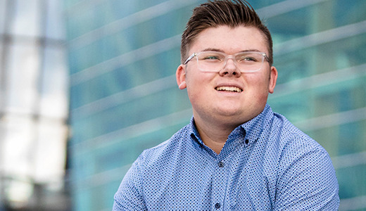 A high school senior with short hair and glasses, wearing a blue patterned shirt, is smiling while looking slightly off-camera. They are outside in front of a modern building with large glass windows.