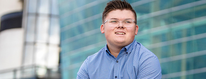 A high school senior with short hair and glasses, wearing a blue patterned shirt, is smiling while looking slightly off-camera. They are outside in front of a modern building with large glass windows.