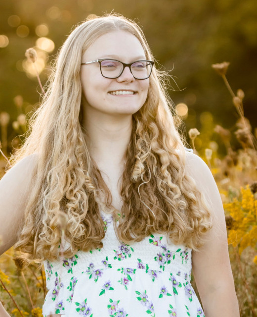 A person with long, curly hair and glasses stands smiling in a flower field of tall yellow blossoms, wearing a white dress adorned with small colorful floral patterns. The blurred greens and yellows of the background hint at a warm, sunny day, perfect for capturing senior pictures.