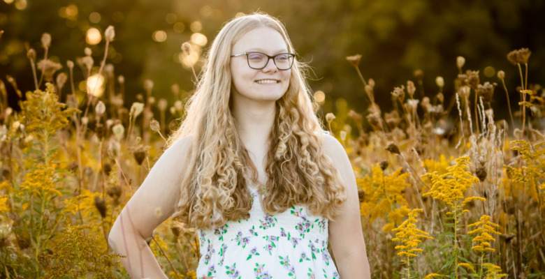 A person with long, curly hair and glasses stands smiling in a flower field of tall yellow blossoms, wearing a white dress adorned with small colorful floral patterns. The blurred greens and yellows of the background hint at a warm, sunny day, perfect for capturing senior pictures.