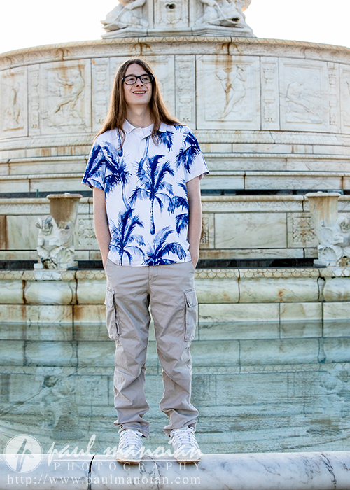 A high school senior with long hair, glasses, and a smile stands in front of a fountain during their senior pictures in Detroit. They are wearing a white shirt with blue palm tree prints, beige cargo pants, and white sneakers. The fountain boasts intricate marble details with water visible at the base.